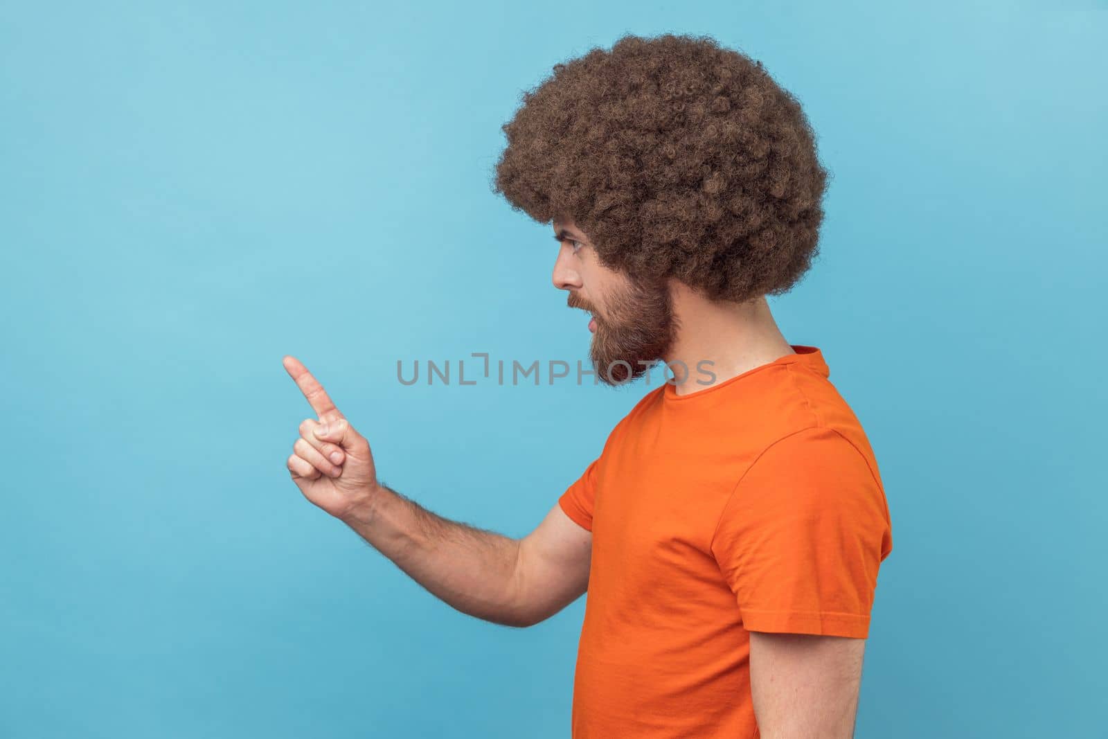 Side view of serious man with Afro hairstyle wearing orange T-shirt standing with admonishing gesture, scolding for mistake and warning. Indoor studio shot isolated on blue background.