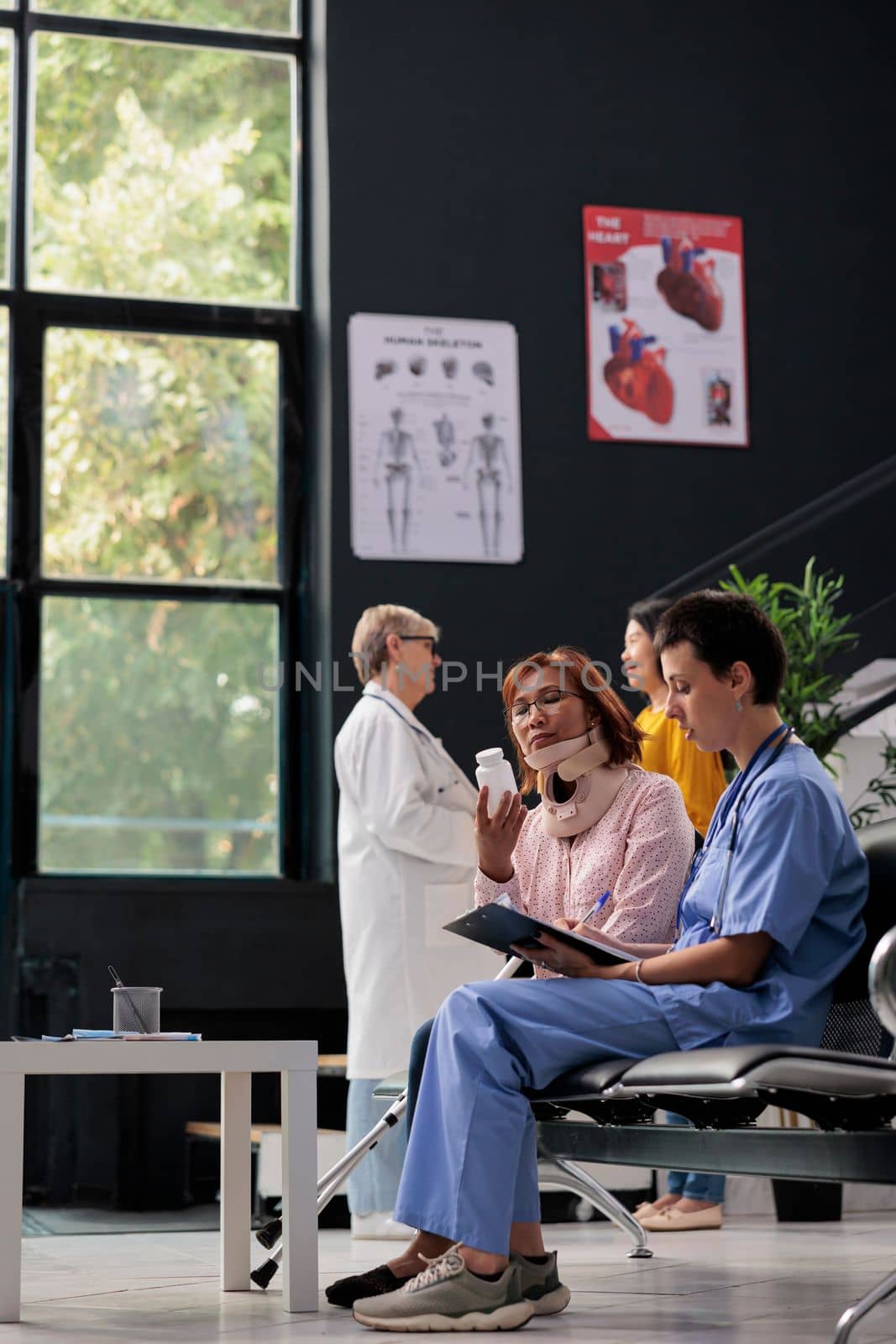 Medical assistant giving bottle pills to injured patient explaining medication treatment during medical appointment. Senior woman wearing cervical neck collar having consultation in hospital lobby