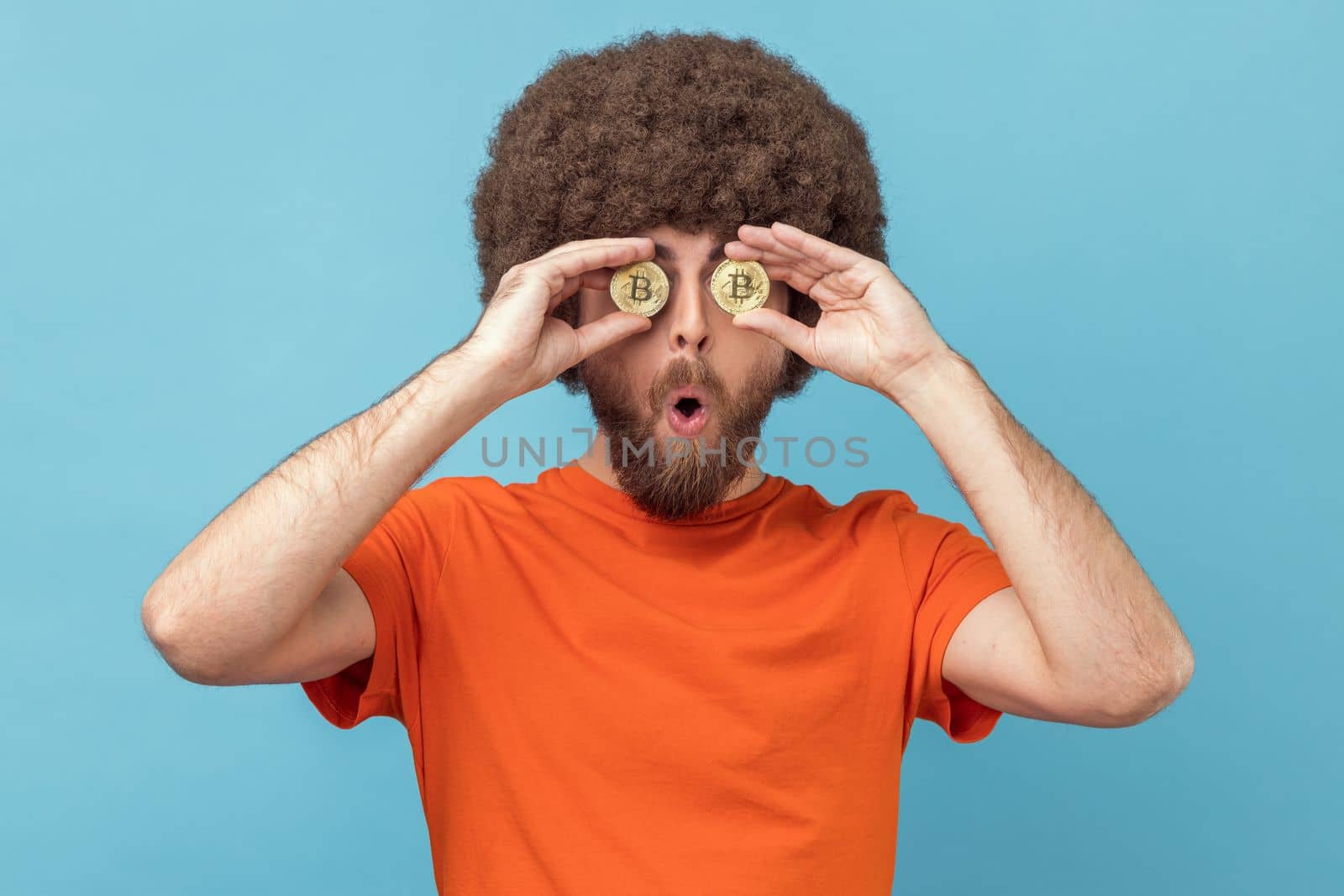 Portrait of astonished man with Afro hairstyle wearing orange T-shirt covering eyes with golden bitcoin, looking at camera through btc coin. Indoor studio shot isolated on blue background.