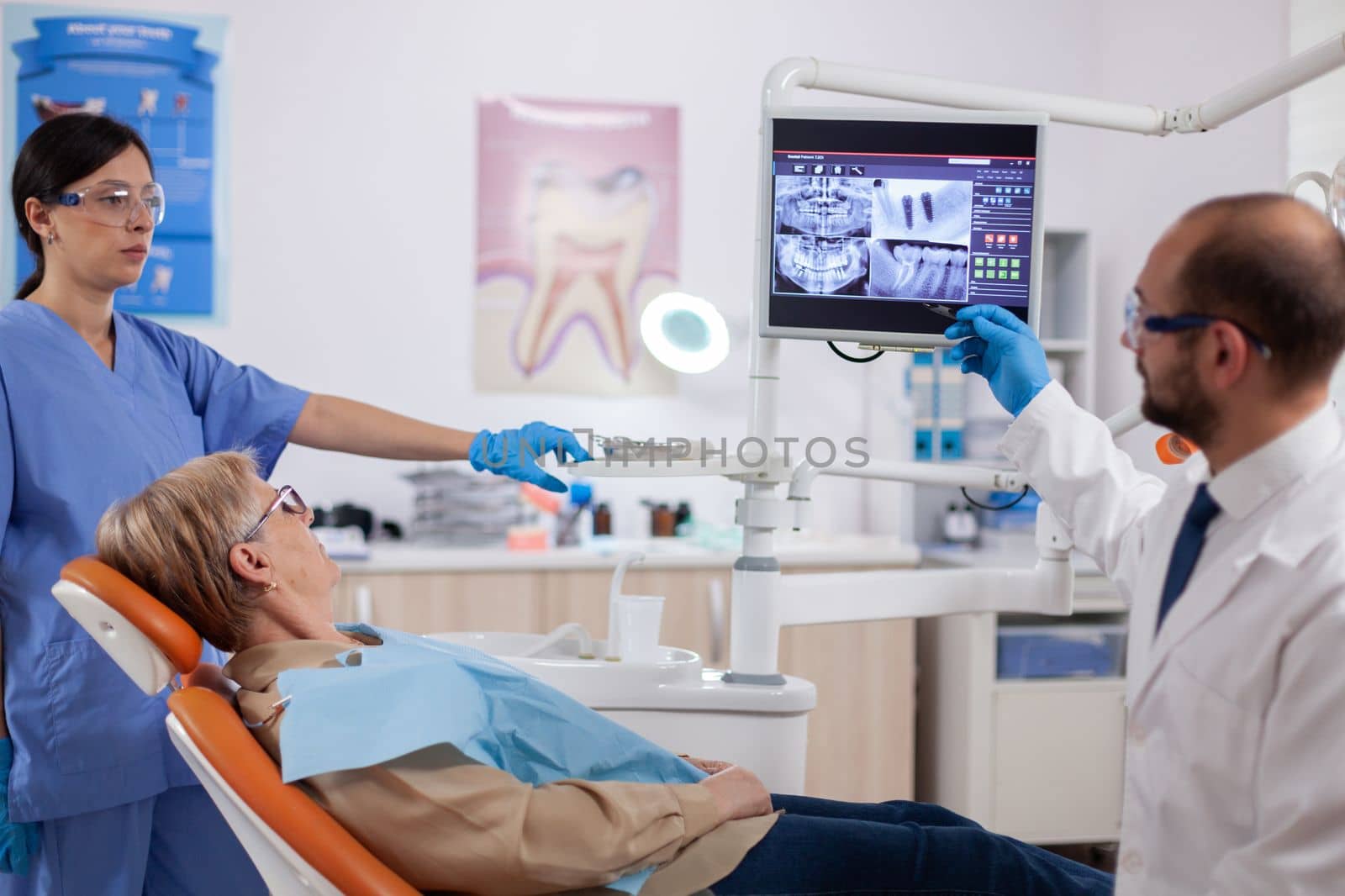 Stomatolog explaining diagnosis to senior woman during dental examination. Medical teeth care taker pointing at patient radiography on screen sitting on chair.