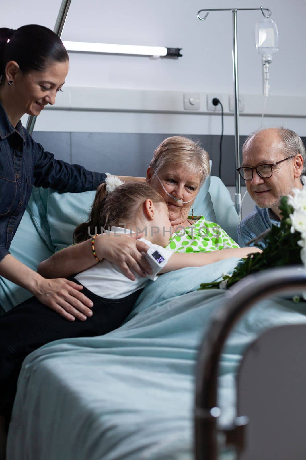 Elderly woman suffering illness lying in hospital observation room bed, hugging young granddaughter during relatives visit. Family giving flowers bouquet to sick grandmother at medical clinic.