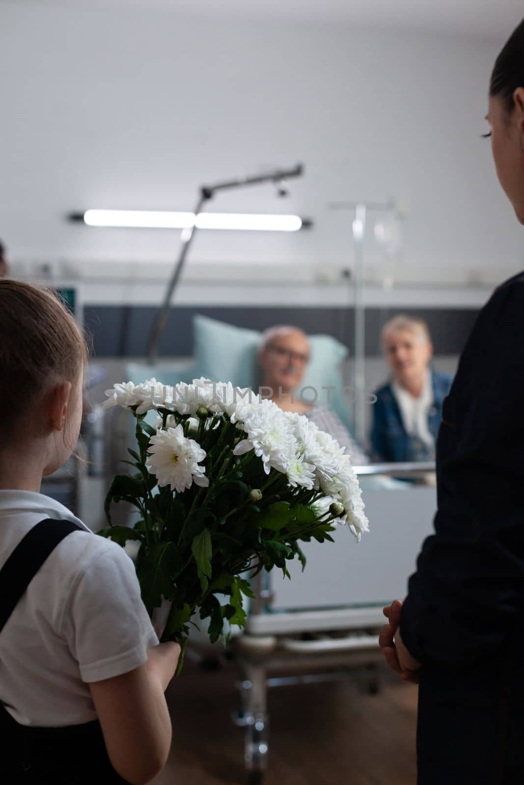 Elderly couple being visited by daughter, granddaughter in geriatric clinic rest room. Little girl carrying flowers gift to recovering grandfather lying in hospital bed.