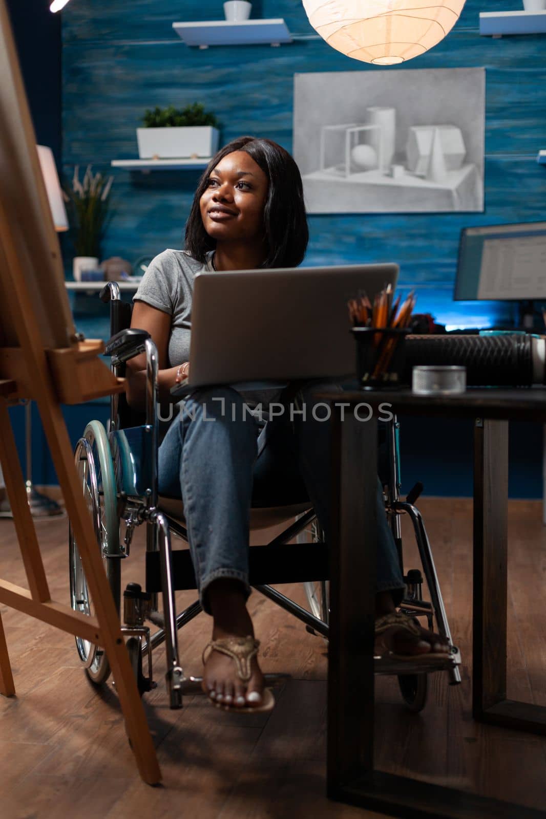 Female sketch artist wheelchair user using laptop computer to take drawing classes remotely at home made studio. African american looking for inspiration online to create artwork.