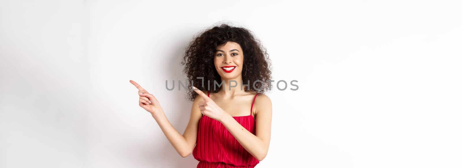 Cheerful female model in fashionable red dress, smiling and pointing fingers right at logo, standing over white background.