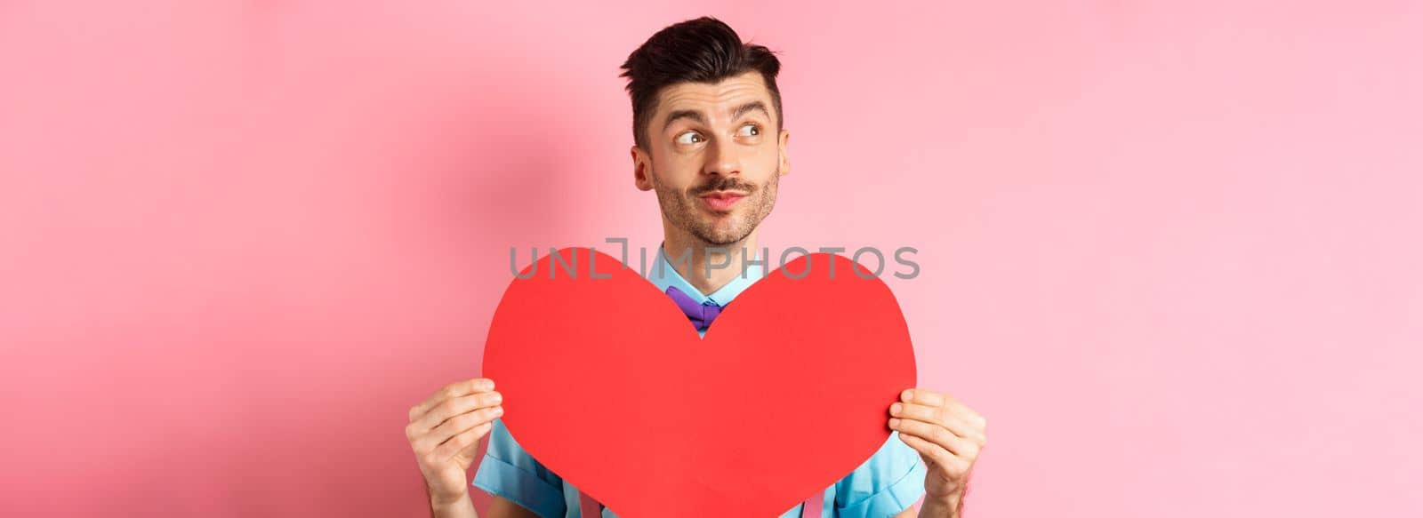 Valentines day concept. Dreamy romantic man looking left and showing big red heart cutout, standing on pink background.