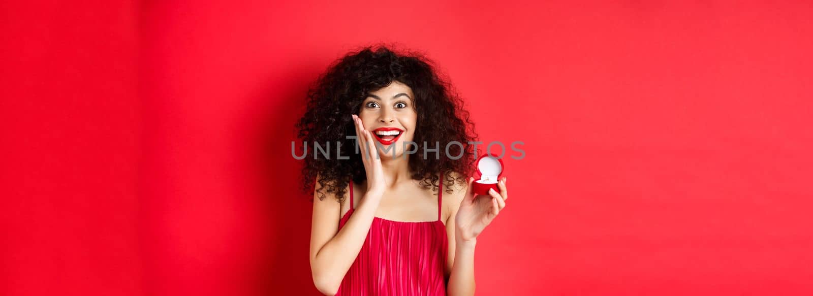 Image of surprised young woman with curly hairstyle, wearing red dress and lipstick, showing engagement ring, going to get married, standing on studio background.