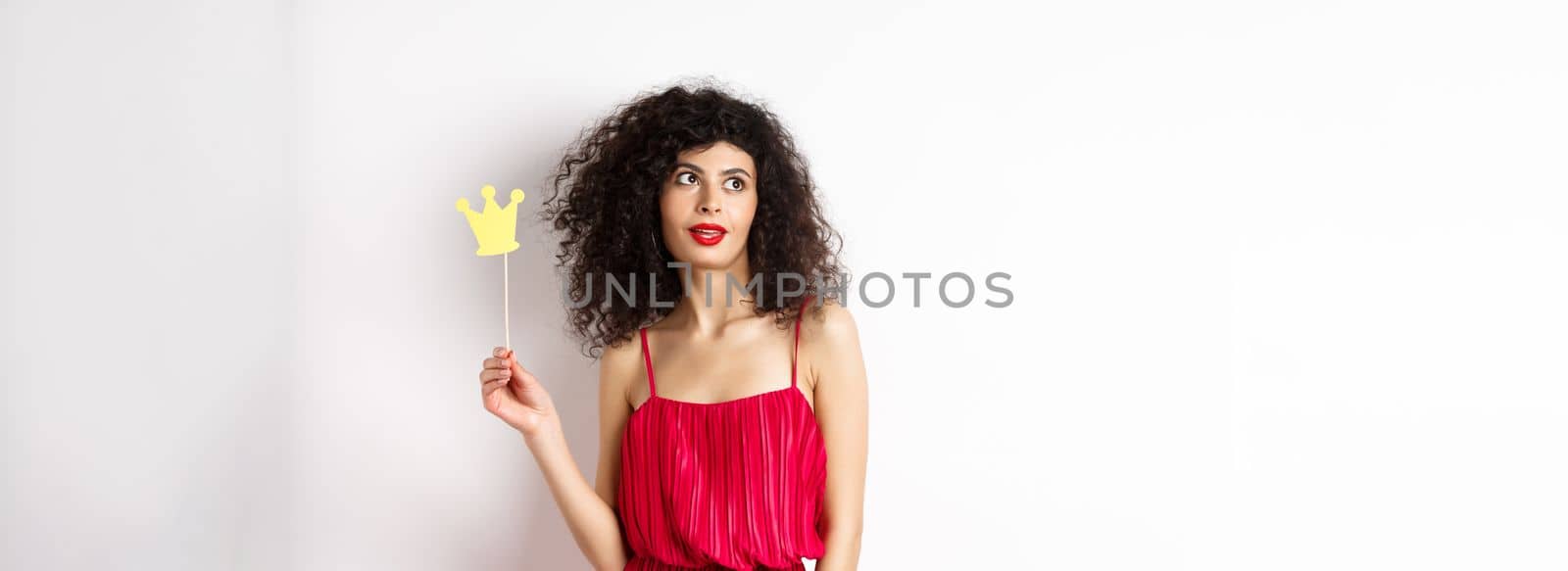 Stylish woman with curly hair in red dress, holding queen crown on stick and looking aside, standing on white background by Benzoix