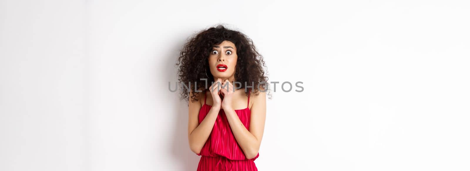 Scared caucasian woman trembling from fear, wearing red dress and staring anxious at camera, standing over white background.