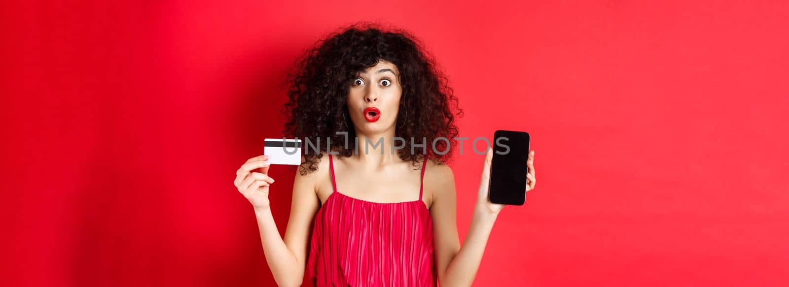 Online shopping concept. Beautiful woman in red dress and lipstick, showing empty phone screen and plastic credit card, standing on studio background.