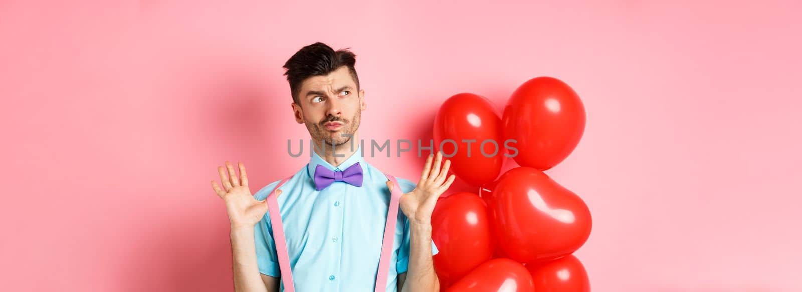 Valentines day concept. Pensive young man in bow-tie, raising hands up and looking left while thinking, making decision, standing on romantic pink background and heart balloons.