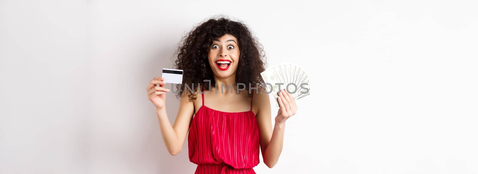 Happy attractive woman in red dress, screaming of joy and showing plastic credit card with money, winning prize, standing over white background by Benzoix