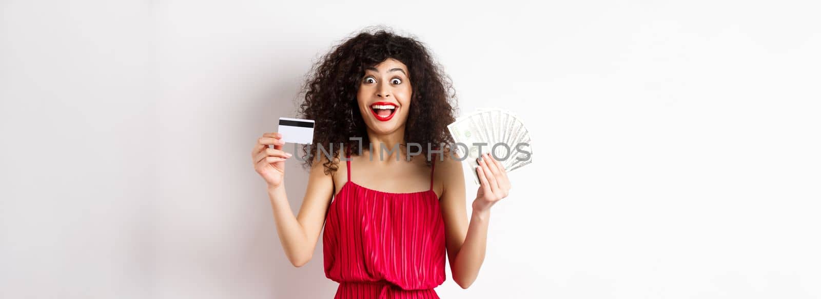 Happy attractive woman in red dress, screaming of joy and showing plastic credit card with money, winning prize, standing over white background.