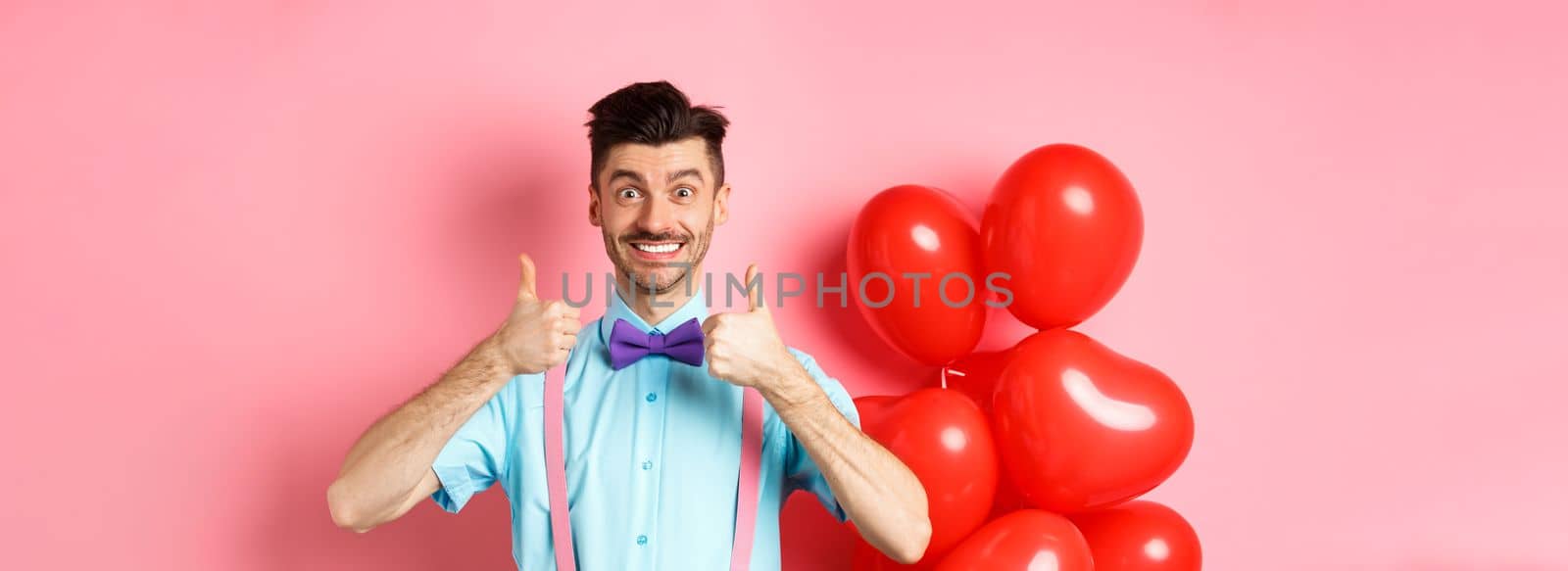 Valentines day concept. Smiling caucasian man showing thumbs up in approval, recommending special deal for lover, standing near cute red hearts balloons and pink background by Benzoix