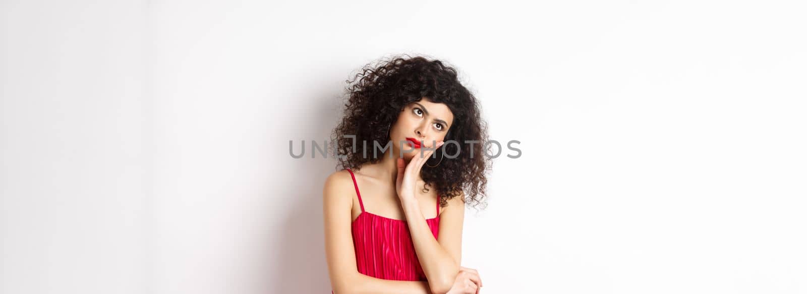 Annoyed and bored young woman with curly hair, look away distressed, lean face on hand, standing bothered in red dress on white background.