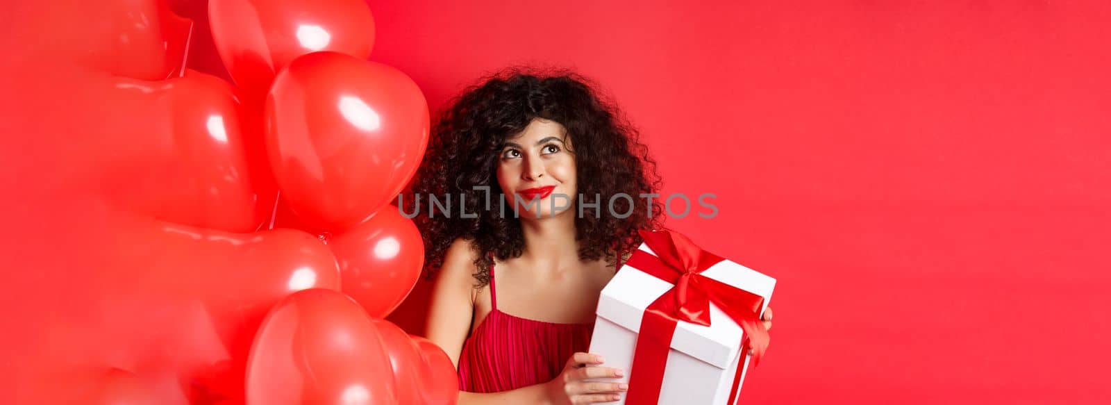 Valentines day and love concept. Beautiful woman with romantic makeup, holding gift box from secret admirer and looking at upper left corner dreamy, standing with heart balloons on red background by Benzoix