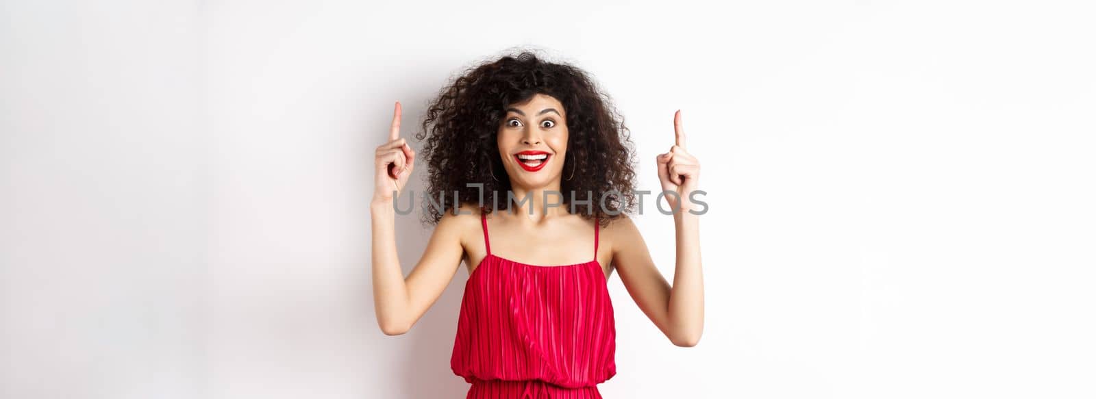 Happy elegant woman in red dress and makeup, smiling amused and pointing fingers up at logo, showing advertisement, standing over white background.
