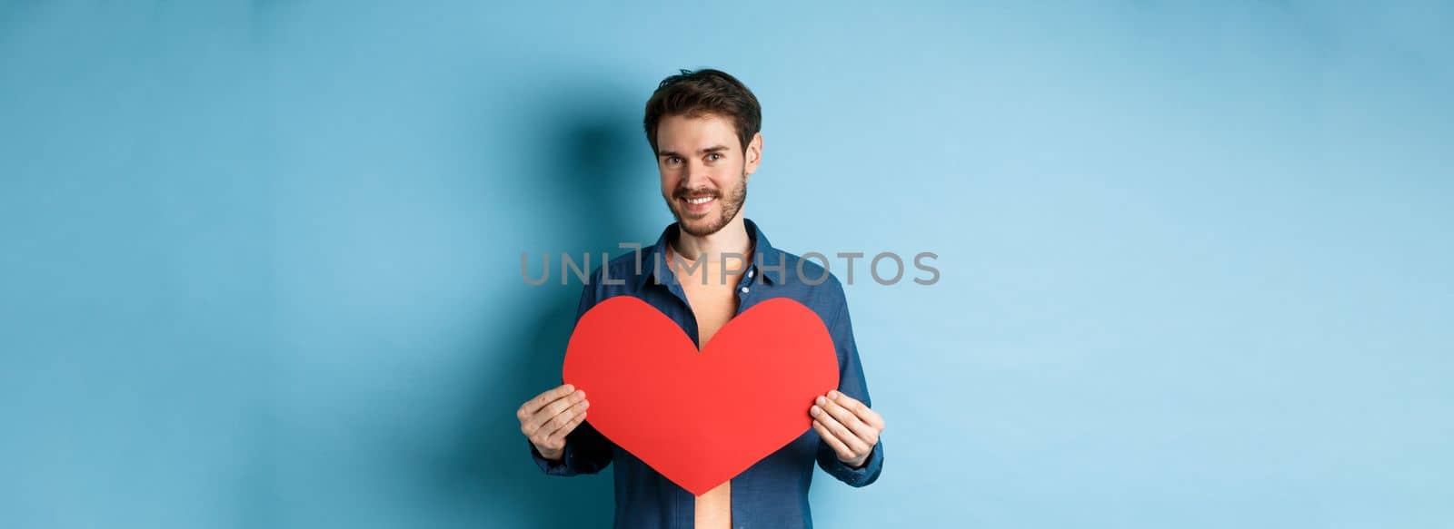 Handsome young man smiling, showing big red heart postcard for Valentines day, looking at camera happy, standing against blue background.