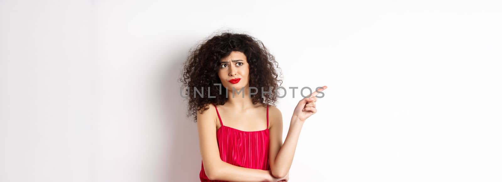 Disappointed and skeptical young woman with curly hair, wearing red dress, grimacing and pointing finger left at logo, standing over white background by Benzoix