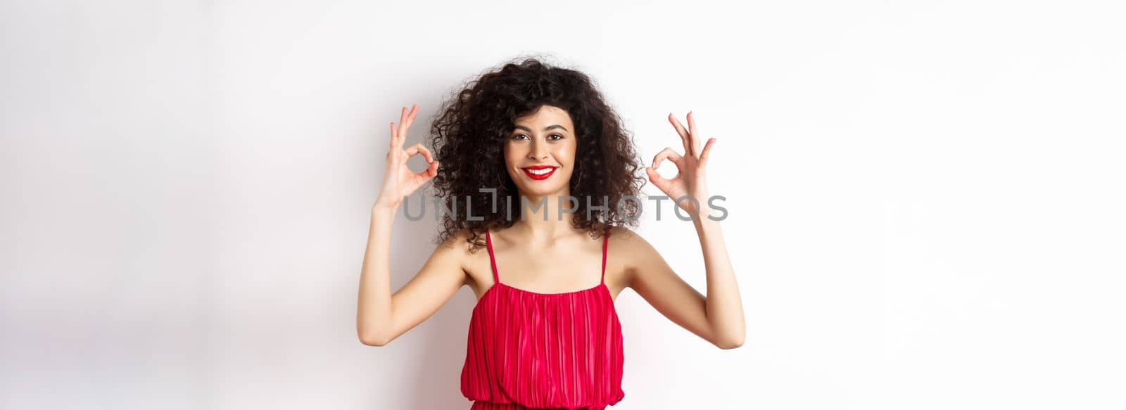 Beautiful woman in red dress, smiling and showing okay gestures, like something good, praising good job, standing over white background by Benzoix