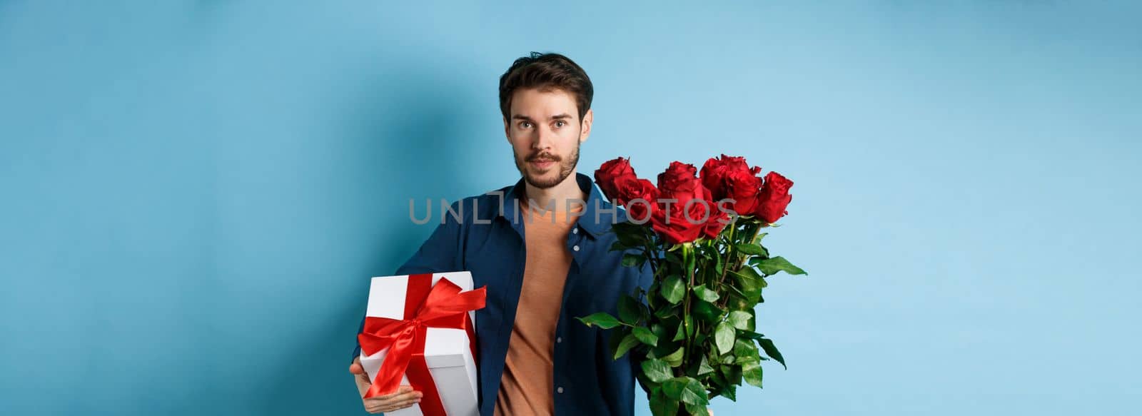 Romance and valentines day. Man presenting bouquet of red roses to lover. Boyfriend bring flowers and gift on romantic date, standing over blue background.