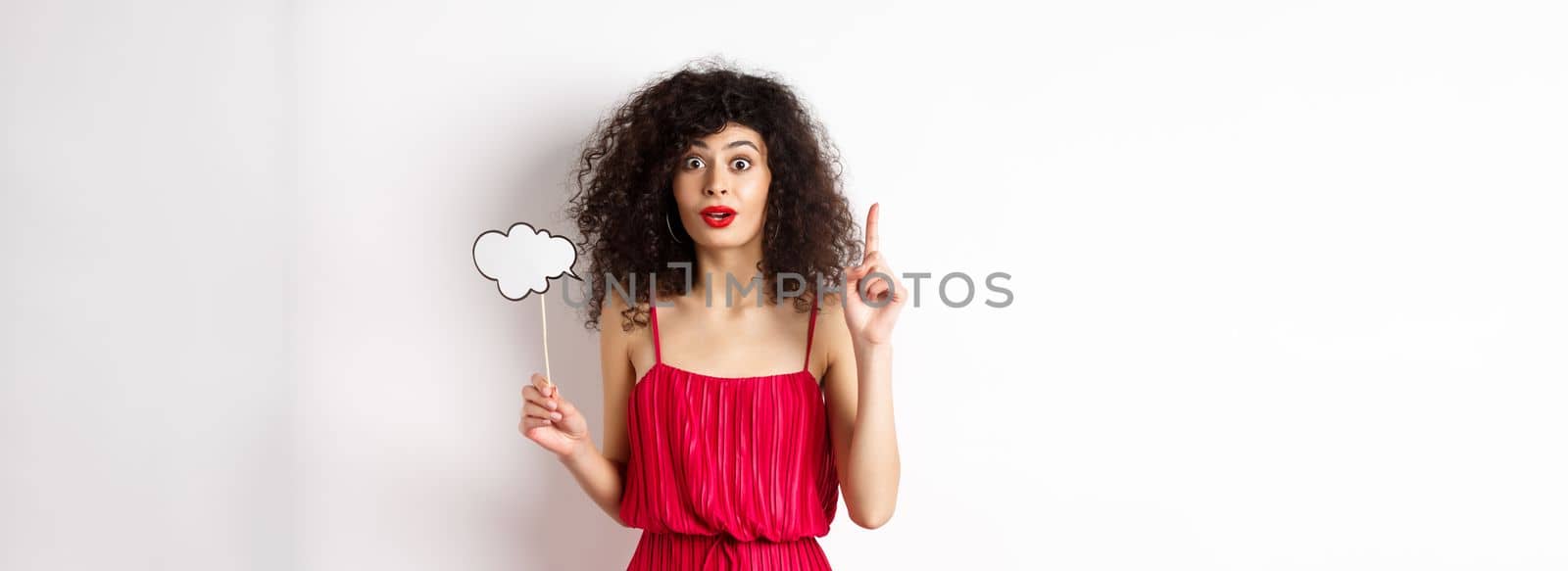 Cute caucasian lady in red dress, holding comment cloud and raising finger, pitching an idea and smiling, suggesting something, standing over white background.