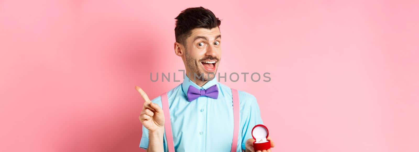 Valentines day. Funny guy with moustache and bow-tie, showing engagement ring and pointing finger at upper right corner, standing on pink background.