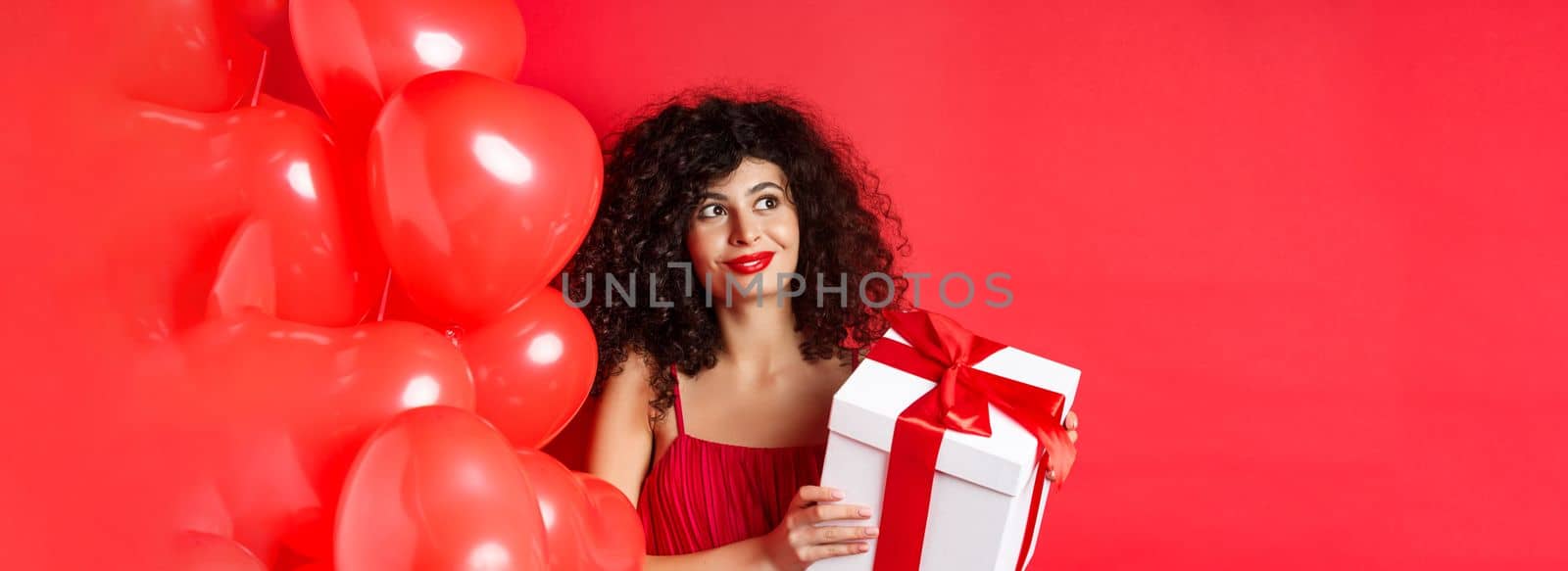 Gorgeous woman with makeup and evening dress, receive surprise gift and smiling, standing on red background near Valentines day romantic balloons.