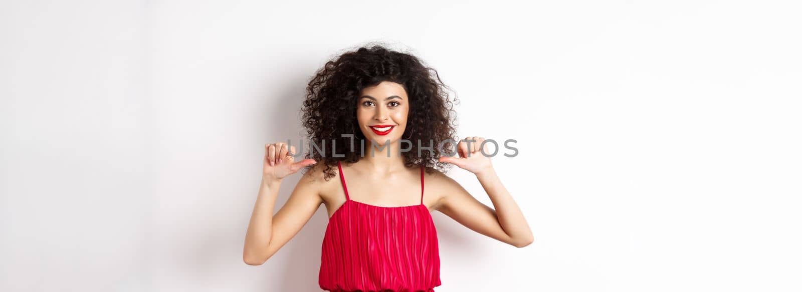 Confident young woman in elegant red dress, pointing at herself and smiling, self-promoting, standing over white background.