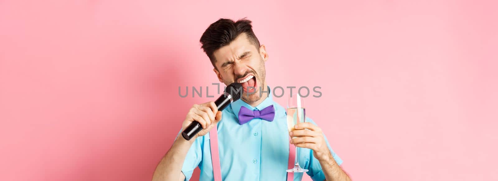 Party and festive events concept. Drunk funny guy singing in microphone and drinking champagne from glass, having fun at karaoke bar, standing on pink background.