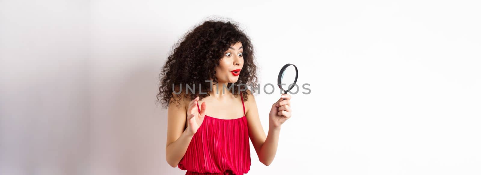 Excited woman in red dress look through magnifying glass and smiling, found interesting promo, standing on white background.