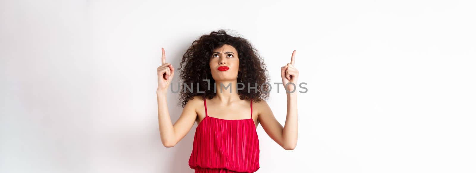 Angry and grumpy woman with curly hair, wearing red dress, frowning and looking up disappointed, standing over white background.