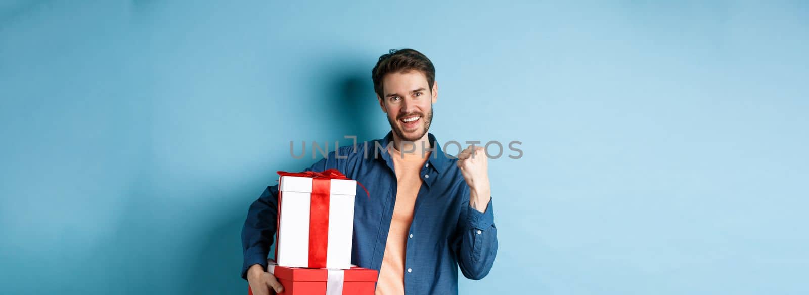 Cheerful young man got discounts on valentines day, making fist pump and say yes, holding gift boxes with presents for lover, standing over blue background by Benzoix