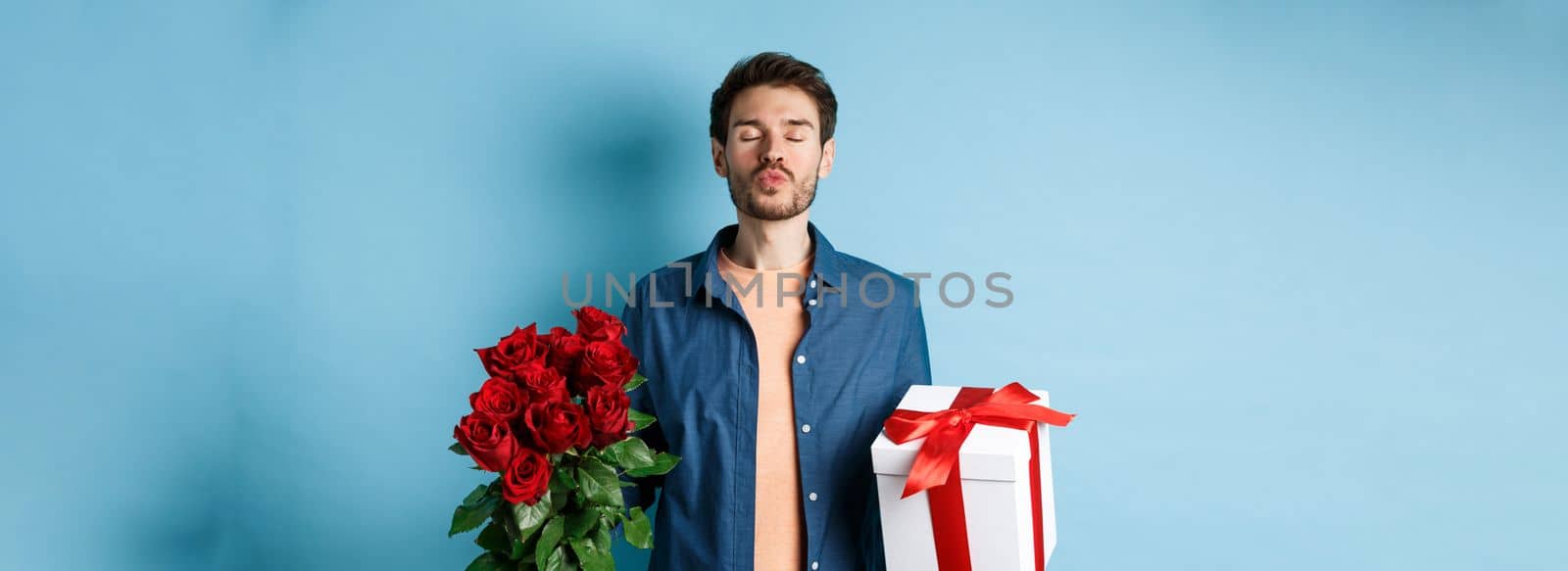 Love and Valentines day concept. Romantic man waiting for kiss, holding gift box and bouquet of red roses for lover on date, standing over blue background by Benzoix