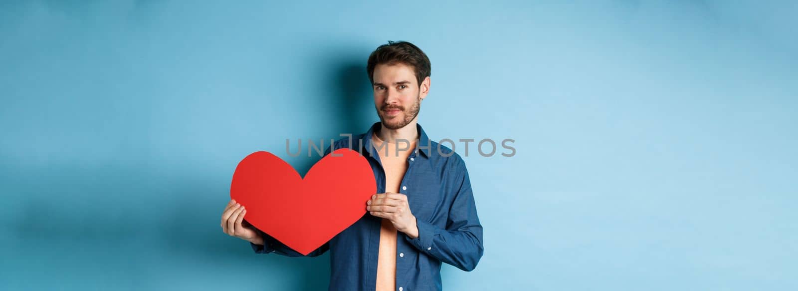 Handsome bearded guy showing valentines day cutout and smiling, standing in casual clothes over blue background by Benzoix
