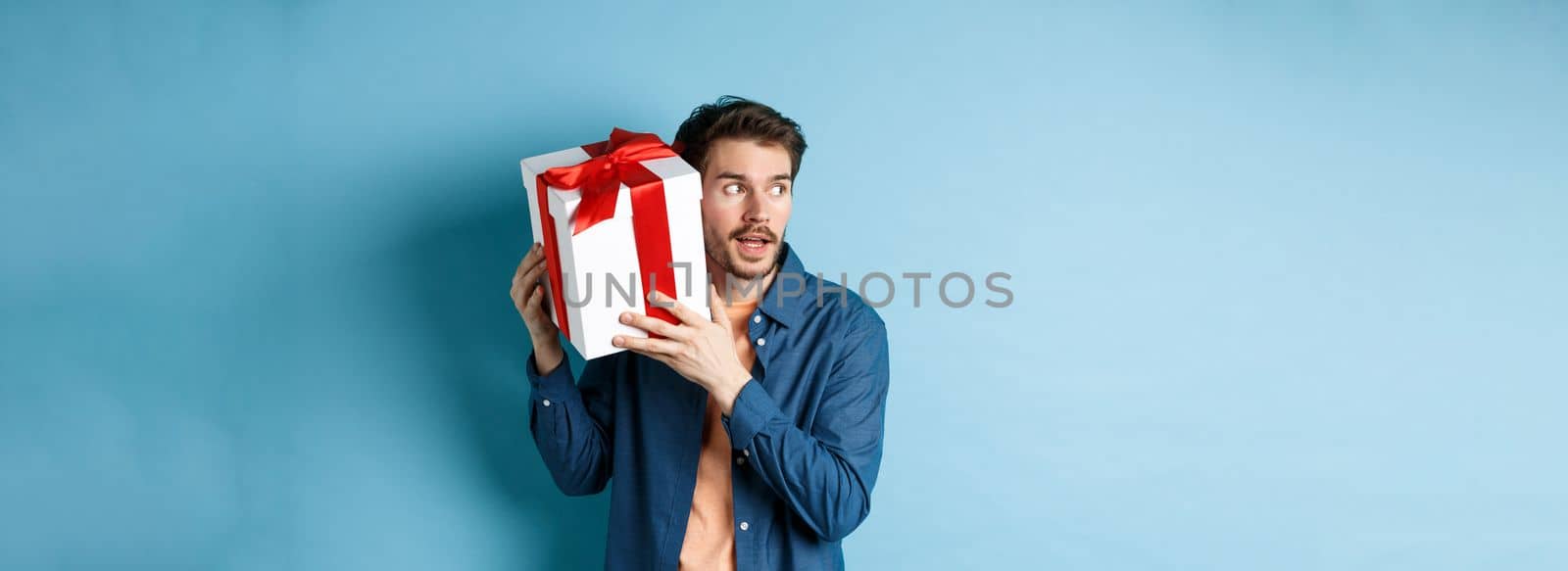 Valentines day. Curious boyfriend holding gift box near ear, trying to guess whats inside surprise present, standing over blue background.