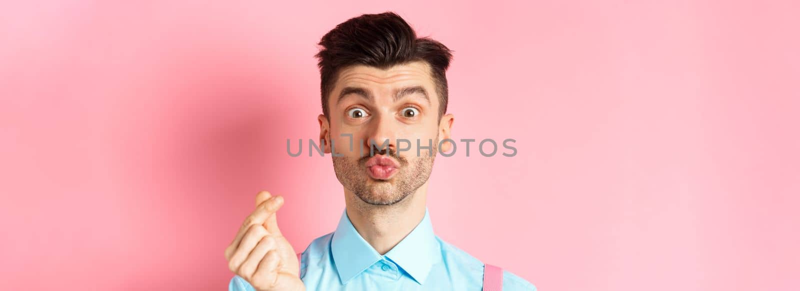 Valentines day concept. Silly guy waiting for kiss and showing finger heart gesture, express love and sympathy, pink background by Benzoix