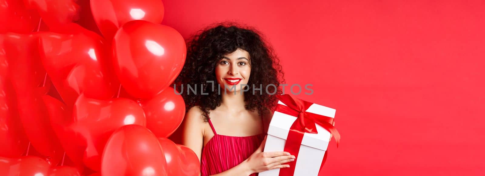 Holidays and celebration. Beautiful woman with curly hair, standing near heart balloons, holding gift box and smiling happy, white background by Benzoix