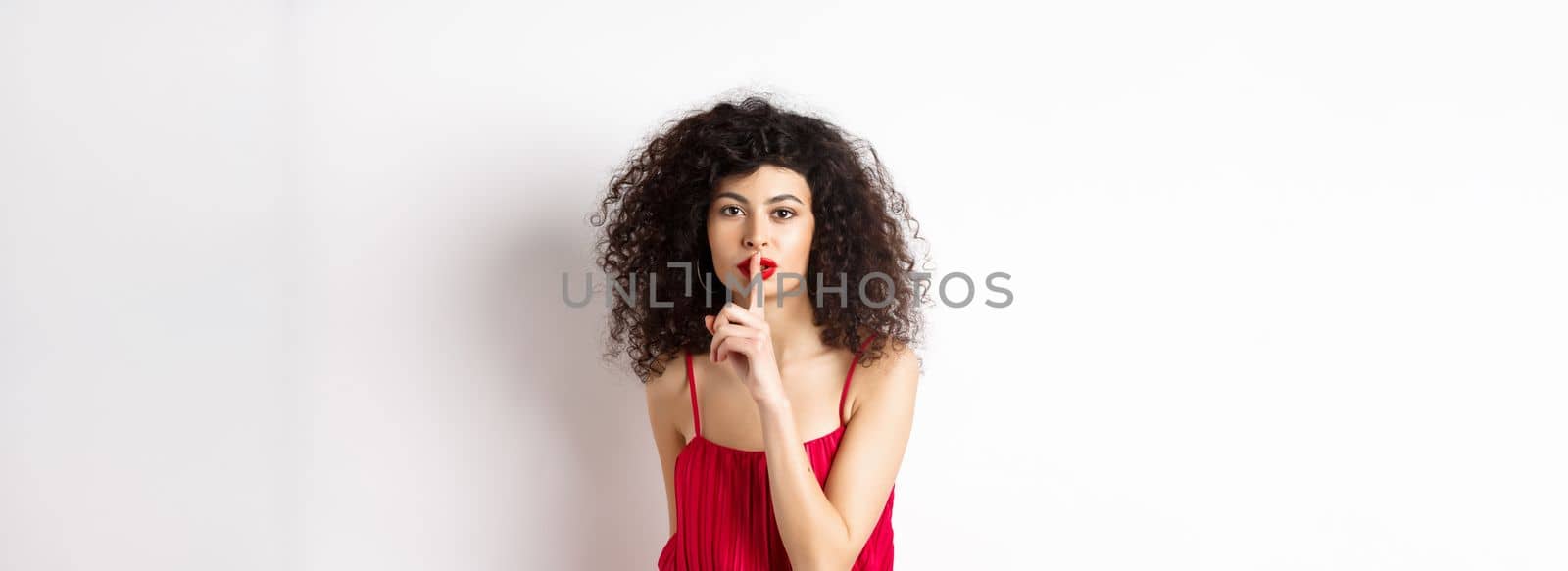 Beautiful woman with curly hair, wearing red dress, hushing at camera, telling secret, shushing at you, standing over white background by Benzoix