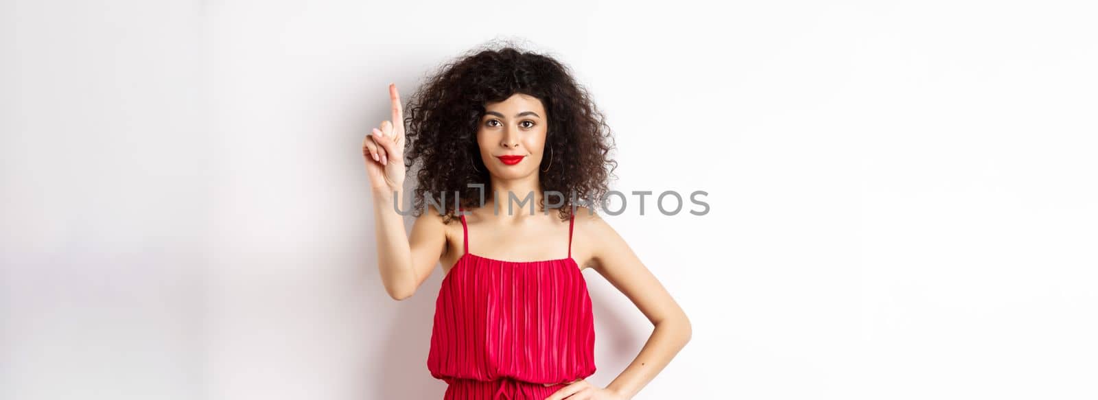 Smiling caucasian woman with curly hair, wearing red dress, showing rule number one gesture, raising finger and looking confident at camera, white background by Benzoix