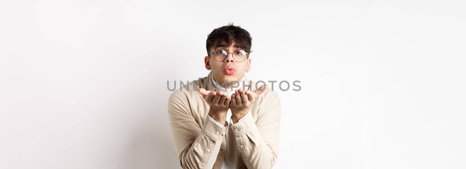 Cute boyfriend sending air kiss at camera on happy Valentines day, looking tender at lover, standing in casual clothes and glasses on white background.