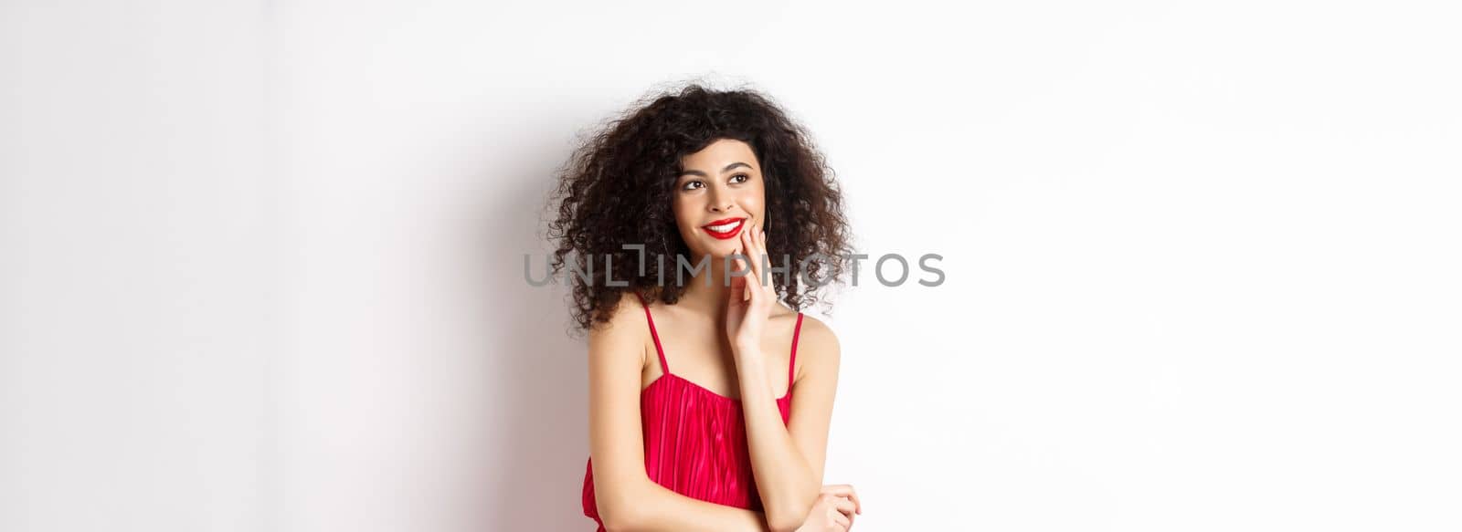 Dreamy elegant girl with curly hair, wearing red dress and makeup, looking left and smiling at logo, standing on white background.