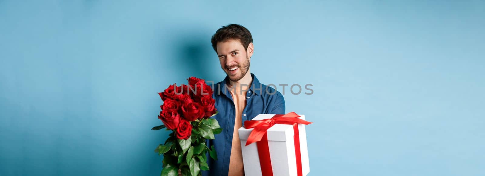 Love and Valentines day concept. Happy guy winking and smiling at camera, presenting a gift and bouquet of flowers to girlfriend, standing over blue background by Benzoix