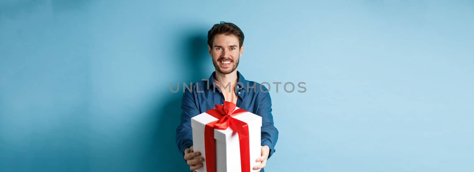 Valentines day. Handsome smiling man extending hands with gift box, wishing happy holiday. Guy making surprise present and looking cheerful, standing over blue background by Benzoix