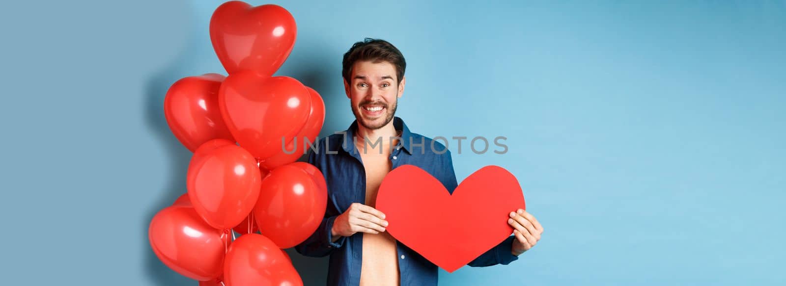 Valentines day concept. Smiling man say I love you, holding paper red heart cutout, standing near romantic balloons, blue background.