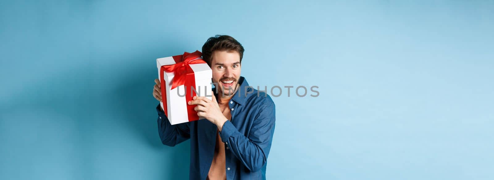 Valentines day. Happy young man got present on special holiday, trying guess what inside gift box and smiling, standing over blue background by Benzoix