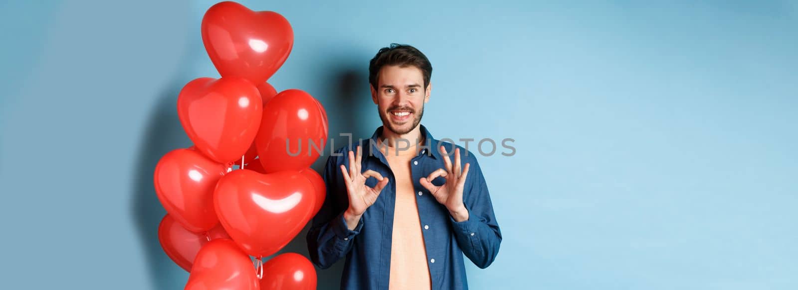 Happy valentines day. Cheerful boyfriend showing okay gestures and praise something good, standing with red hearts balloons for lover, blue background by Benzoix