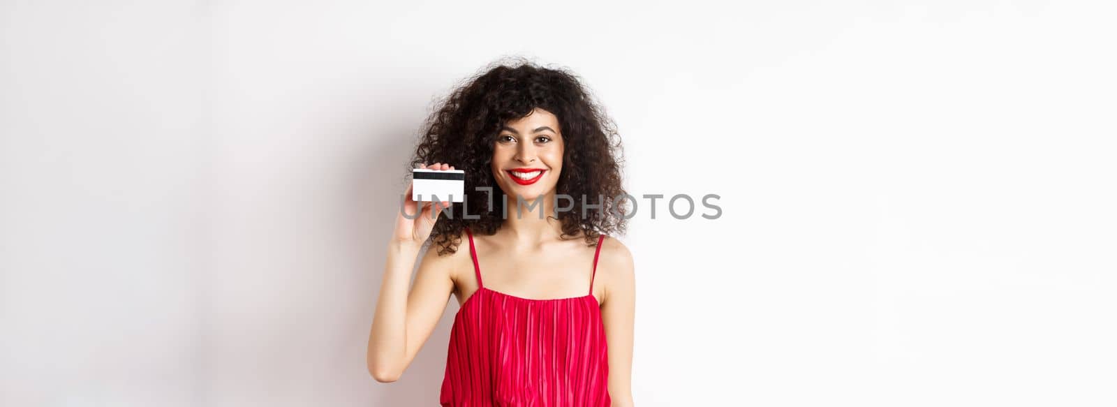 Beautiful woman in red trendy dress and makeup, showing plastic credit card and smiling, recommending offer, standing on white background.