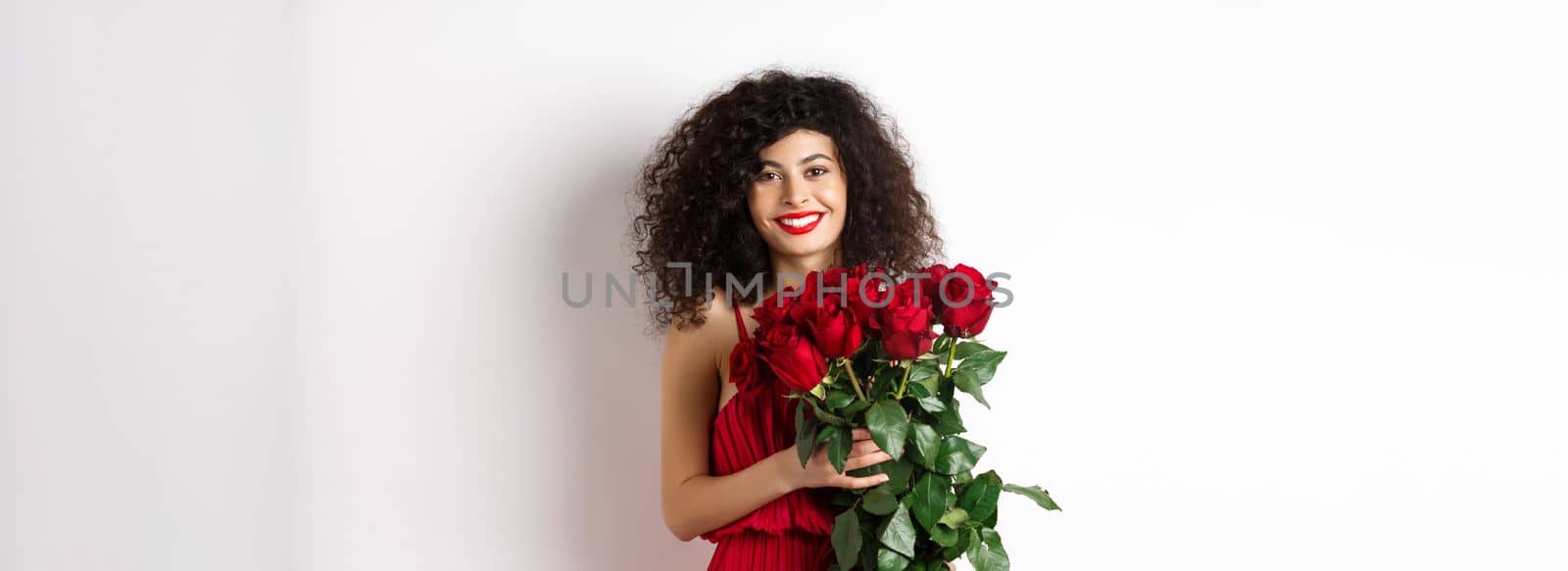 Gorgeous caucasian woman in red evening dress receiving bouquet of flowers. Girl with red roses smiling at camera, having romantic date on Valentines lover day, white background by Benzoix