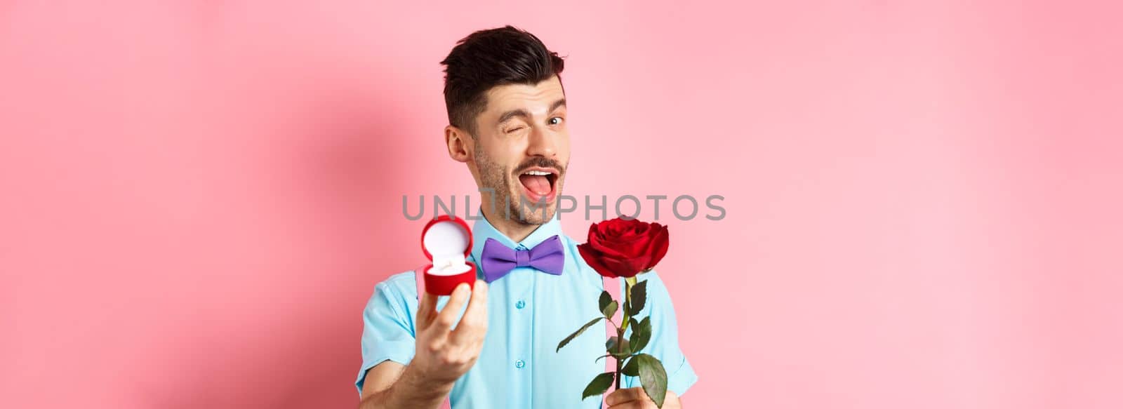 Valentines day. Funny guy making proposal, winking and saying marry me, showing engagement ring with red rose, standing over pink background.