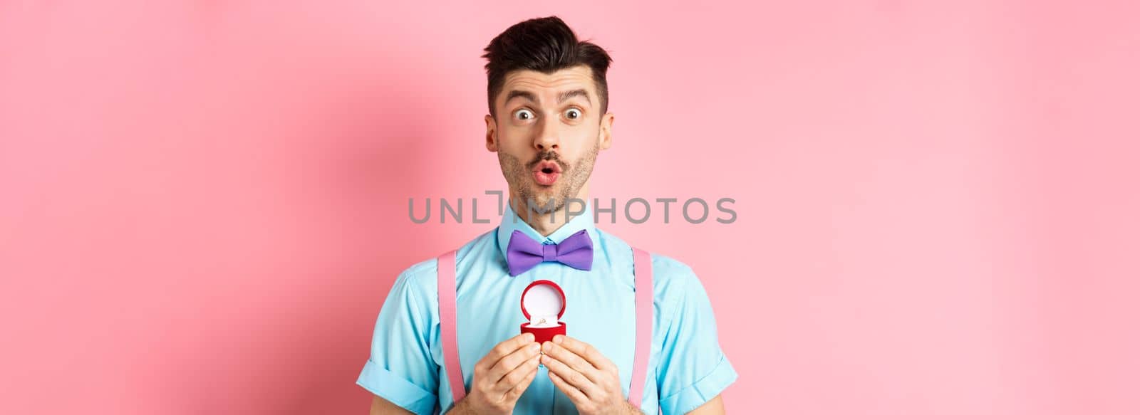 Valentines day. Funny man with moustache and bow-tie, looking excited and showing engagement ring, asking girlfriend to marry him, standing over pink background.